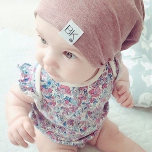 K&ouml;nnte beinhalten: Ein Baby tr&auml;gt eine rosa M&uuml;tze mit einem wei&szlig;en Etikett, auf dem "BK co." steht, und einen Strampler mit Blumenmuster.