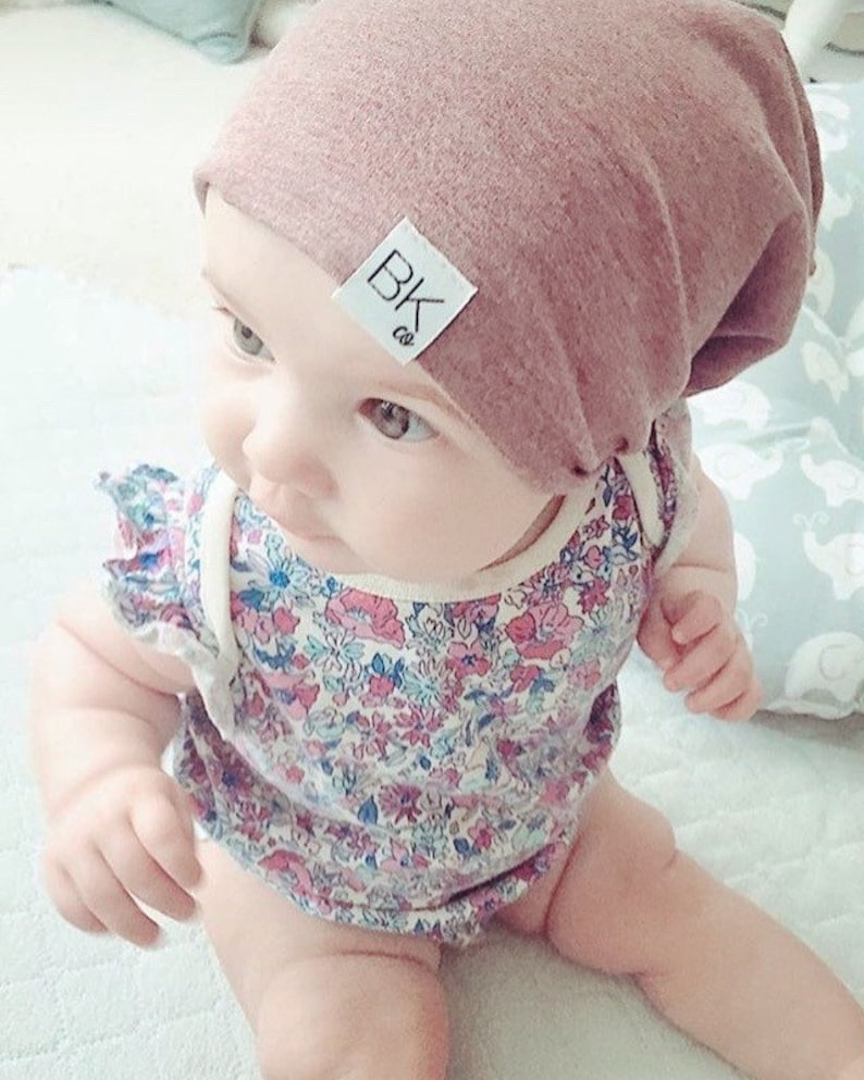 K&ouml;nnte beinhalten: Ein Baby tr&auml;gt eine rosa M&uuml;tze mit einem wei&szlig;en Etikett, auf dem "BK co." steht, und einen Strampler mit Blumenmuster.