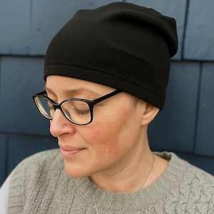 May include: A black beanie hat. The hat is made of a soft material and is worn by a person wearing glasses and a grey sweater. The background is a blue wall.