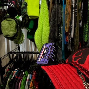 May include: A display of various apparel items, including hats, beanies, and clothing. A purple cassette tape pin is in the foreground, with a red hat featuring a flame design. The background shows a rack of colorful clothing.