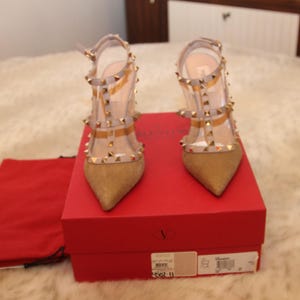 Pu&ograve; includere: Un paio di scarpe col tacco alto dorate con cinturini trasparenti e borchie dorate. Le scarpe sono esposte su una scatola rossa con un sacchetto rosso.