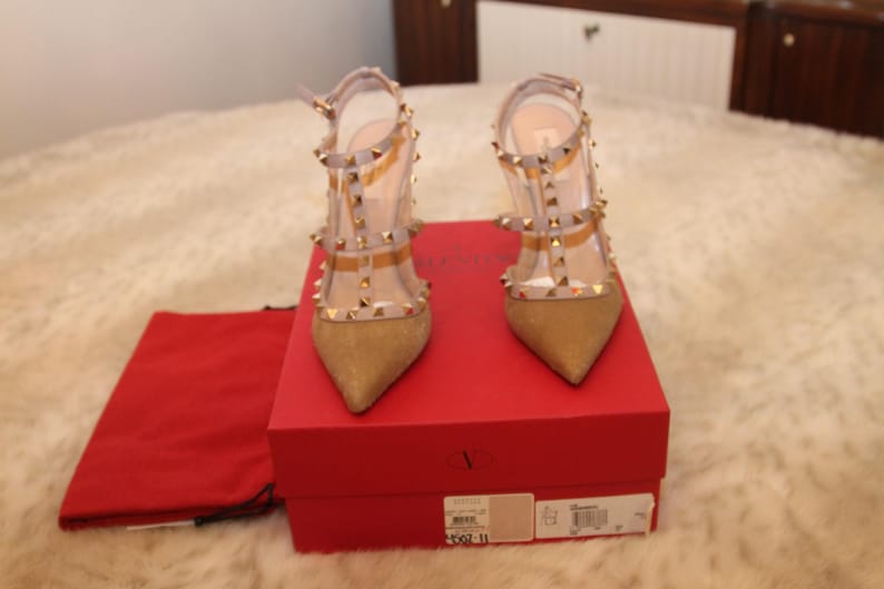 Pu&ograve; includere: Un paio di scarpe col tacco alto dorate con cinturini trasparenti e borchie dorate. Le scarpe sono esposte su una scatola rossa con un sacchetto rosso.