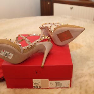 Pu&ograve; includere: D&eacute;collet&eacute; in camoscio beige con borchie dorate e sottili cinturini. Le scarpe sono esposte su una scatola rossa con il logo del marchio. I tacchi hanno la punta e un tacco a spillo. La scatola misura 25 cm.