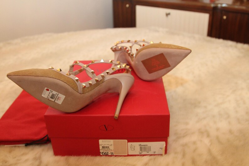 Pu&ograve; includere: D&eacute;collet&eacute; in camoscio beige con borchie dorate e sottili cinturini. Le scarpe sono esposte su una scatola rossa con il logo del marchio. I tacchi hanno la punta e un tacco a spillo. La scatola misura 25 cm.