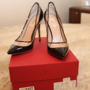 Puede incluir: Un par de zapatos de tac&oacute;n alto de charol negro con paneles transparentes, sobre una caja roja. Los zapatos tienen punta afilada y tacones de aguja. La caja tiene una etiqueta con n&uacute;meros y texto.