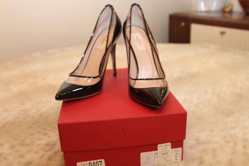 Puede incluir: Un par de zapatos de tac&oacute;n alto de charol negro con paneles transparentes, sobre una caja roja. Los zapatos tienen punta afilada y tacones de aguja. La caja tiene una etiqueta con n&uacute;meros y texto.
