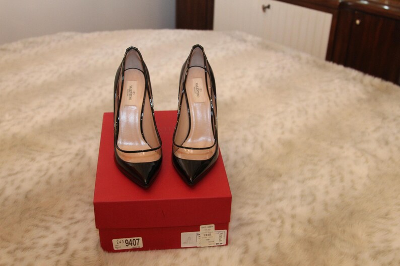 Puede incluir: Un par de zapatos de tac&oacute;n alto de charol negro con detalles de PVC transparente, expuestos sobre una caja roja. Los zapatos tienen punta afilada y la marca es visible. La caja tiene una etiqueta con n&uacute;meros.