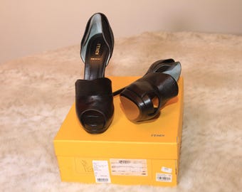 Op de afbeelding: Zwarte leren open-teen high heels met een peep-toe design. De schoenen staan op een gele Fendi doos. De hakken hebben een plateauzool en een dunne enkelband. De binnenkant van de schoen is lichtblauw.