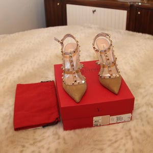 Pu&ograve; includere: Un paio di scarpe col tacco alto in camoscio beige e PVC trasparente con borchie dorate. Le scarpe sono esposte su una scatola rossa, accanto a un sacchetto rosso con coulisse. Le scarpe hanno la punta e cinturini alla caviglia.