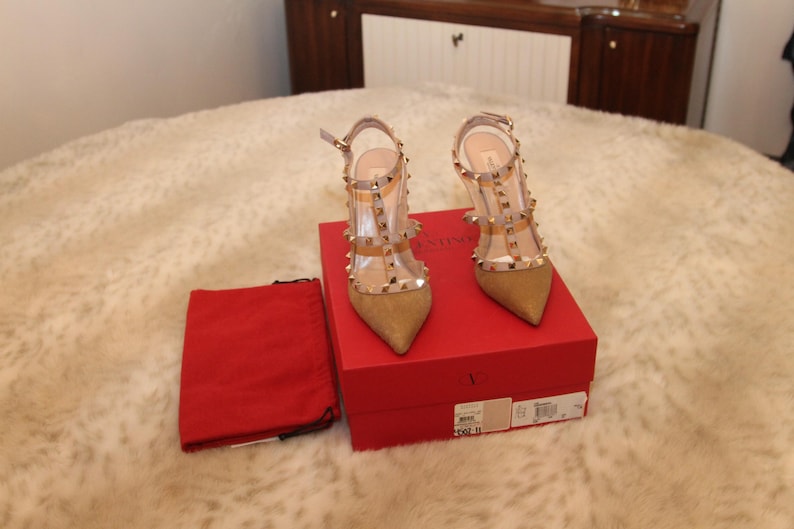 Pu&ograve; includere: Un paio di scarpe col tacco alto in camoscio beige e PVC trasparente con borchie dorate. Le scarpe sono esposte su una scatola rossa, accanto a un sacchetto rosso con coulisse. Le scarpe hanno la punta e cinturini alla caviglia.