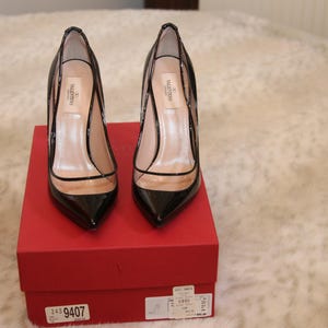 Puede incluir: Un par de zapatos de tac&oacute;n alto de charol negro con detalles de vinilo transparente, expuestos sobre una caja roja. Los zapatos tienen punta afilada y tac&oacute;n de aguja. La caja tiene una etiqueta del producto.