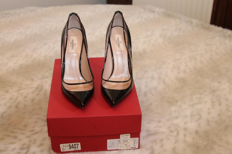 Puede incluir: Un par de zapatos de tac&oacute;n alto de charol negro con detalles de vinilo transparente, expuestos sobre una caja roja. Los zapatos tienen punta afilada y tac&oacute;n de aguja. La caja tiene una etiqueta del producto.