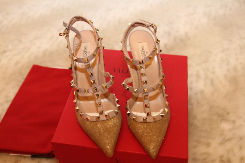 Pu&ograve; includere: Un paio di d&eacute;collet&eacute; dorate con glitter e punta a punta, con pi&ugrave; cinturini e borchie dorate. Le scarpe sono su una scatola rossa con un sacchetto rosso. I tacchi sono a spillo.