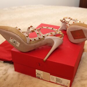 Pu&ograve; includere: Un paio di sandali beige con tacco alto, borchie e cinturini dorati. Le scarpe sono su una scatola rossa con un sacchetto antipolvere rosso abbinato. I tacchi sono sottili e a punta.