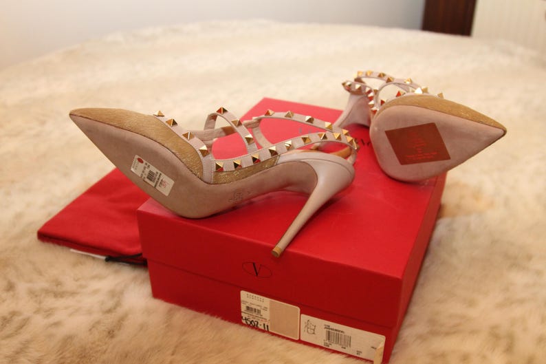 Pu&ograve; includere: Un paio di sandali beige con tacco alto, borchie e cinturini dorati. Le scarpe sono su una scatola rossa con un sacchetto antipolvere rosso abbinato. I tacchi sono sottili e a punta.