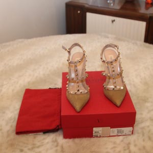 Pu&ograve; includere: Un paio di scarpe col tacco alto color oro con cinturini trasparenti e borchie color oro. Le scarpe sono esposte su una scatola rossa con un sacchetto rosso. I tacchi sono alti circa 10 cm.