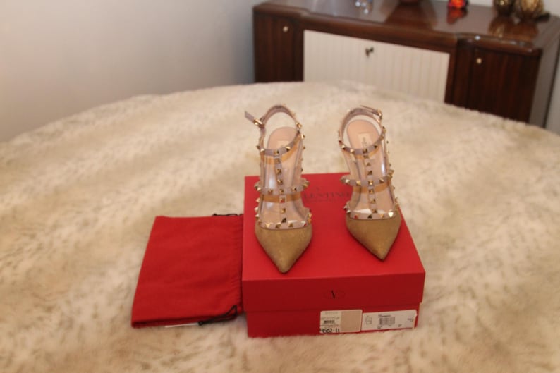 Pu&ograve; includere: Un paio di scarpe col tacco alto color oro con cinturini trasparenti e borchie color oro. Le scarpe sono esposte su una scatola rossa con un sacchetto rosso. I tacchi sono alti circa 10 cm.