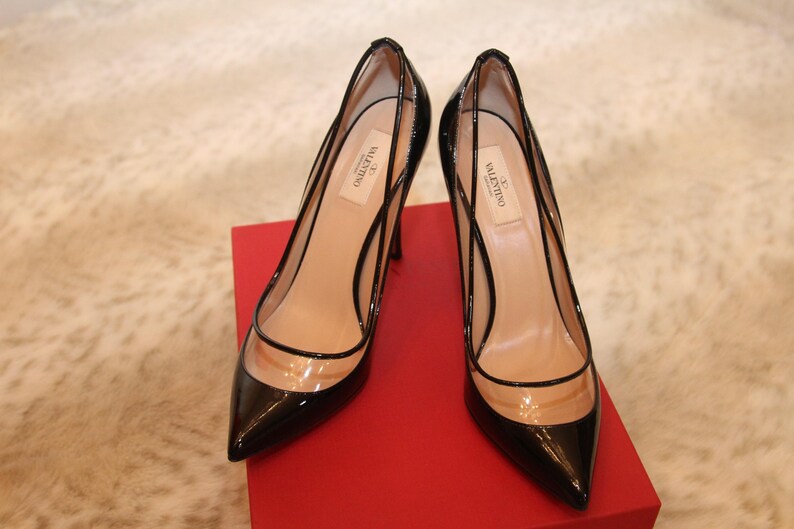 Puede incluir: Un par de zapatos de tac&oacute;n alto de charol negro con paneles de vinilo transparente. Los zapatos tienen punta afilada y se exhiben sobre una caja roja. La marca "Valentino" es visible en la plantilla.