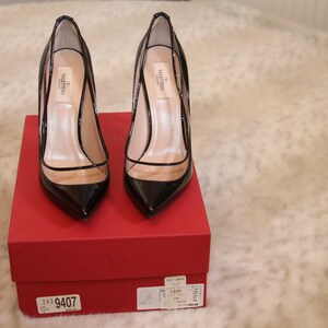 Puede incluir: Un par de zapatos de tac&oacute;n negros de charol con punta afilada y paneles de PVC transparentes, expuestos sobre una caja roja. Los tacones tienen un dise&ntilde;o de aguja. Los zapatos son de la marca Valentino.