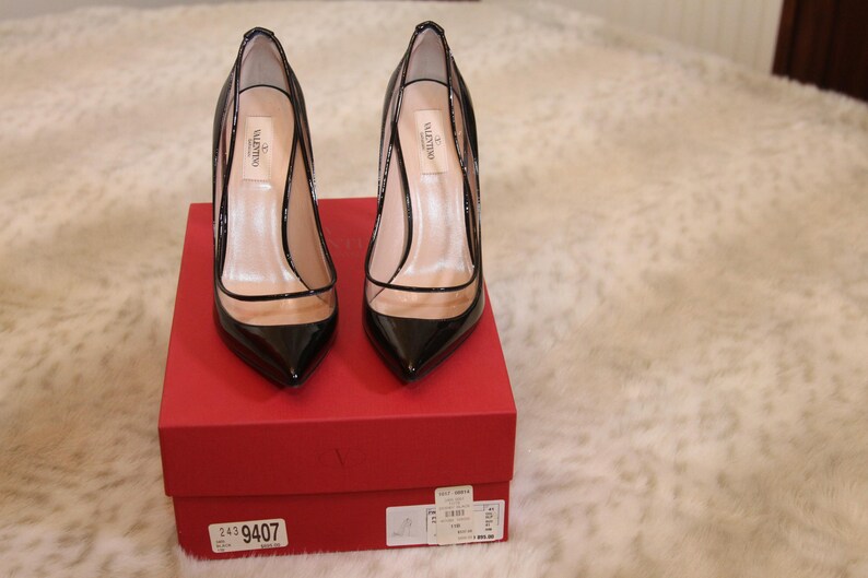 Puede incluir: Un par de zapatos de tac&oacute;n negros de charol con punta afilada y paneles de PVC transparentes, expuestos sobre una caja roja. Los tacones tienen un dise&ntilde;o de aguja. Los zapatos son de la marca Valentino.
