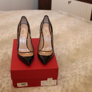 Puede incluir: Un par de zapatos de tac&oacute;n alto de charol negro con punta y paneles transparentes, expuestos sobre una caja roja. Los zapatos tienen tac&oacute;n de aguja. El nombre de la marca es visible.