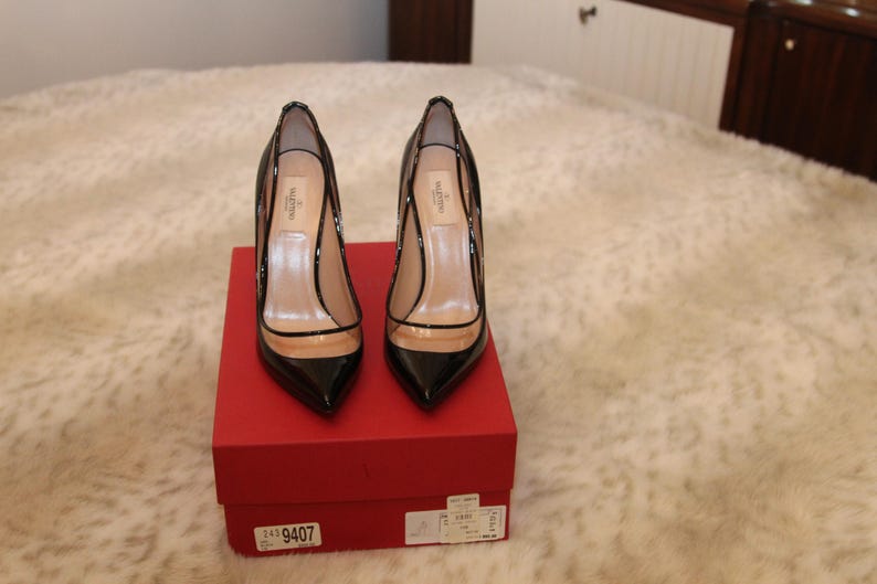 Puede incluir: Un par de zapatos de tac&oacute;n alto de charol negro con punta y paneles transparentes, expuestos sobre una caja roja. Los zapatos tienen tac&oacute;n de aguja. El nombre de la marca es visible.
