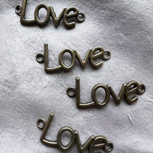May include: Four bronze-colored metal charms spelling the word "Love" in a cursive font. Each charm has small loops on either end for attaching to other items. The charms are arranged on a light gray, textured fabric.