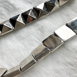 May include: Close-up of two silver metal necklaces. One necklace features pyramid-shaped links, while the other has rectangular links. The jewelry is displayed on a textured, off-white surface.