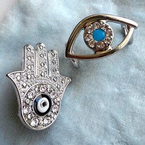 May include: Two silver-toned rings with decorative designs. One ring is shaped like an eye with a turquoise center and clear stones. The other ring is a hand shape with a central eye design and clear stones.