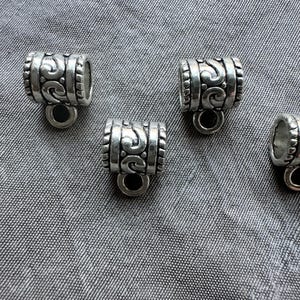 May include: Close-up of several silver-colored metal bead connectors. Each connector has a cylindrical shape with decorative scrollwork and a small loop at the bottom. The beads are arranged on a gray fabric surface.