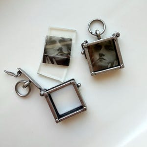 May include: Three antique silver-toned pendants with small square frames. One pendant is open, revealing a blank white space. The other two pendants contain a black and white image of a person's face. A small glass slide with a similar image is also present.