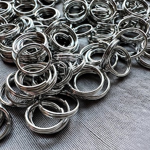 May include: A collection of shiny, silver-colored split key rings. The rings are tightly coiled and arranged in a pile on a textured gray surface. The key rings are made of metal and have a circular shape.
