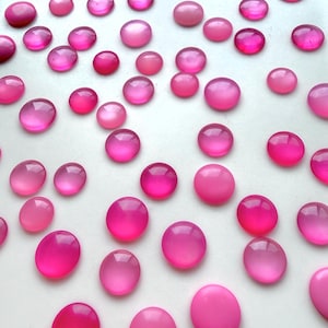 May include: A collection of numerous round, smooth, cabochon-style buttons in various shades of pink, ranging from light pastel to deep magenta. Each button features a polished, glossy finish and a domed top, scattered across a plain white surface.