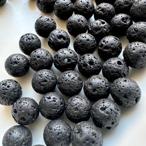 Puede incluir: Primer plano de numerosas cuentas de roca volcánica negra, cada una con una textura porosa distintiva. Las cuentas esféricas se muestran sobre un fondo blanco, resaltando su color oscuro y superficie táctil, ideal para manualidades.