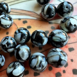 May include: A collection of round beads with a marbled design. The beads are primarily black and white, with swirling patterns creating a unique appearance. The beads are scattered on a patterned surface.