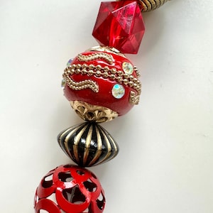 May include: A close-up of a beaded necklace with various decorative elements. The necklace features red, gold, and black beads, including a faceted red gem and a filigree bead. The beads are strung together, creating a striking visual.
