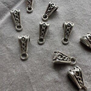 May include: Close-up of several silver-colored pendant bails with an ornate design. Each bail has a loop at the bottom for attaching to a chain or cord. The bails are scattered on a gray fabric surface.