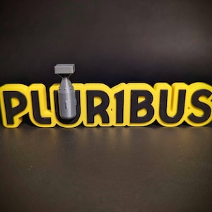 May include: A yellow and black 3D-printed sign that reads "PLURIBUS" with a gray bomb on top. The sign is on a black surface.