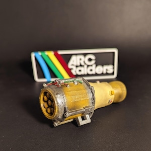 May include: A yellow and silver cylindrical object with a series of holes on one end and a threaded connector on the other. The object has wires and a small rectangular sign with the text "ARC Raiders".
