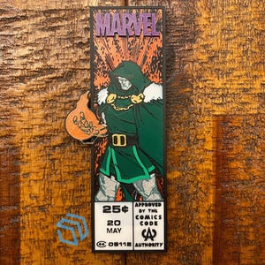 May include: A rectangular Marvel comic book cover bookmark featuring Doctor Doom in a green hooded cloak and metallic armor. The design includes the text MARVEL, 25 cents, 20 MAY, and the Comics Code Authority seal. An orange hand icon extends from the side.