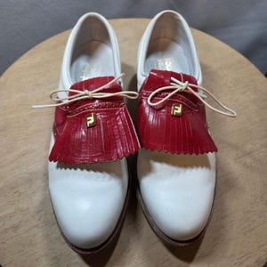May include: A pair of white leather golf shoes with red fringed accents and a gold logo. The shoes have a classic design with a lace-up closure and brown soles. The red accents add a pop of color to the otherwise neutral design.