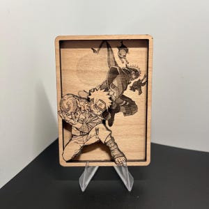 May include: A wooden plaque featuring a detailed laser-etched illustration of anime characters in action poses. The artwork is set against a light wood grain background, mounted on a clear plastic stand.