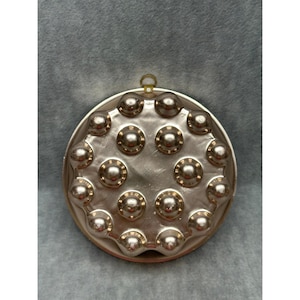 May include: A round, metallic copper-toned gelatin mold featuring a series of raised, circular indentations arranged in a circular pattern. The mold has a smooth, reflective surface and includes a small metal loop at the top for hanging purposes.