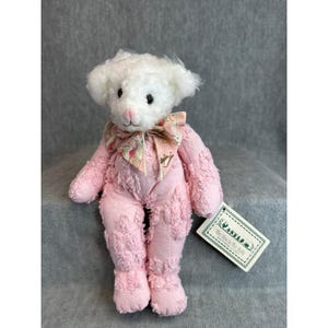 May include: A handmade teddy bear with a white head and pink body. The bear has a pink bow and a tag attached. The bear is wearing a pink outfit with textured details. The bear is sitting on a gray surface.