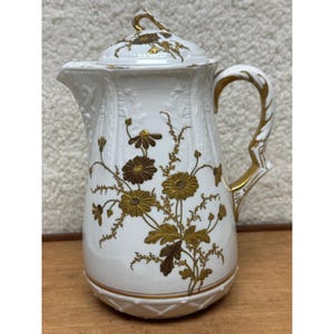May include: A white porcelain coffee pot featuring a gold floral pattern of daisies and foliage. The pot has a matching lid with a gold-trimmed handle, a curved spout, and embossed vertical detailing along the body and base with gold accents.