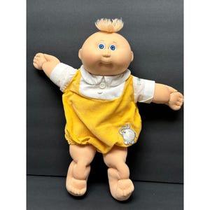 May include: A doll with a round head, light skin, blue eyes, and a small tuft of blonde hair on top. The doll wears a white collared shirt under a yellow corduroy romper featuring a small embroidered gray elephant on the lower right side.