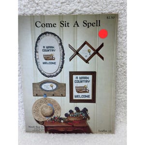 May include: A craft pattern leaflet titled Come Sit A Spell featuring cross-stitch designs of porch swings and straw hats. The cover displays an oval framed sampler, a wooden coat rack, a rectangular framed sampler, and a straw hat with blue ribbon.