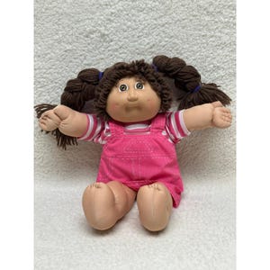 May include: A doll with a soft fabric body, tan skin, and brown yarn hair styled in two pigtails tied with blue bands. The doll wears a pink overall dress with white stitching over a pink and white striped short-sleeved shirt with a round neckline.