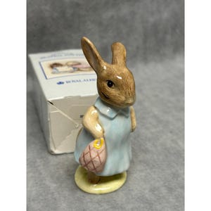 May include: A ceramic figurine of a brown rabbit wearing a light blue dress and holding a pink bag. The figurine is on a yellow base, with a box in the background. The figurine is a collectible item.