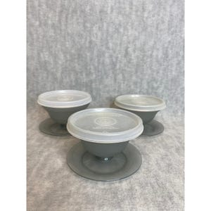 May include: Three gray plastic bowls with clear lids and matching saucers. The bowls have a rounded shape and sit on a small, flat base. The lids are circular and fit snugly on top of the bowls. The set is designed for food storage.
